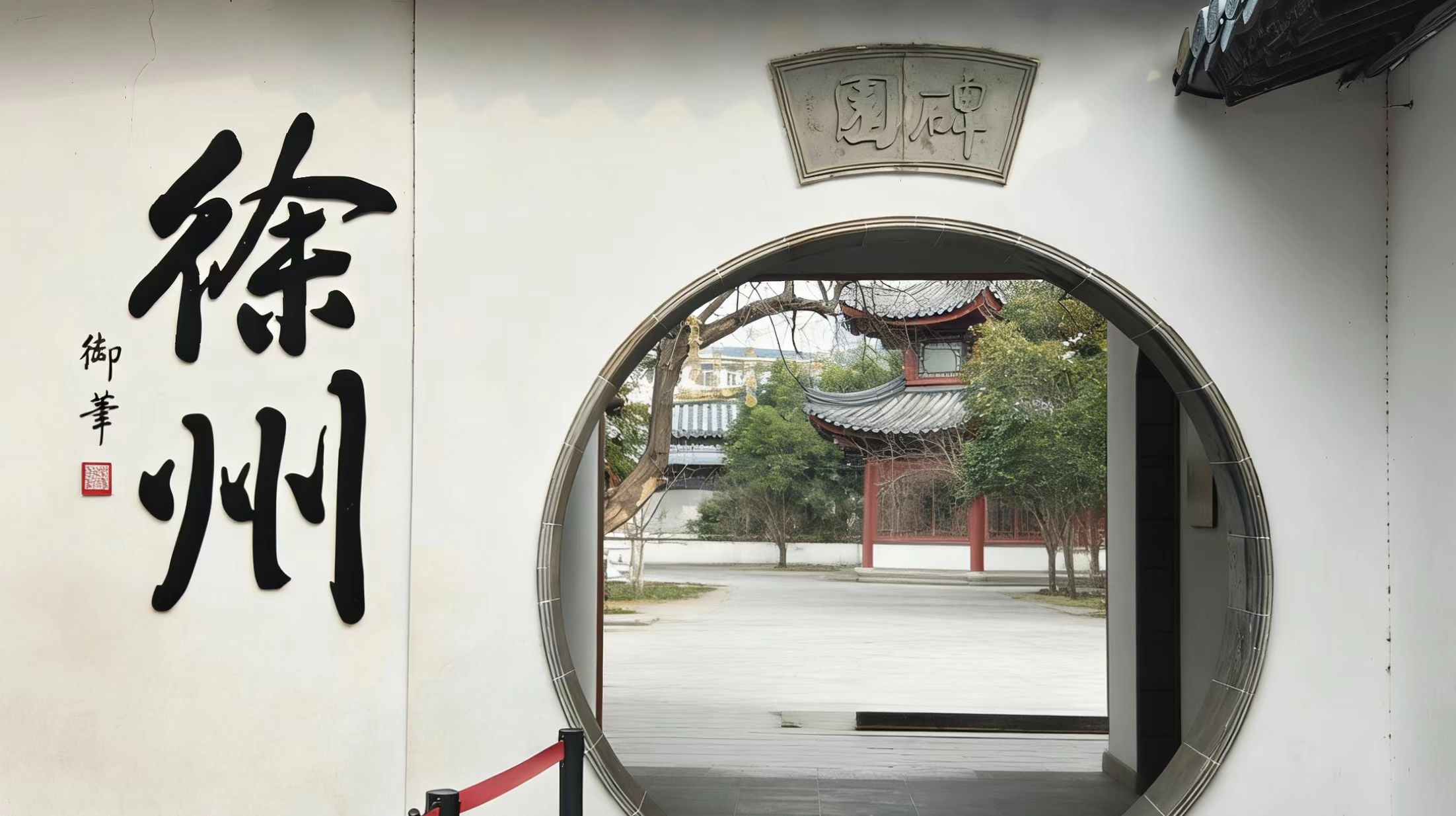 白墙上的圆形月洞门,门上方扇形石刻牌匾,墙侧竖写徐州二字,门内可见亭台树木