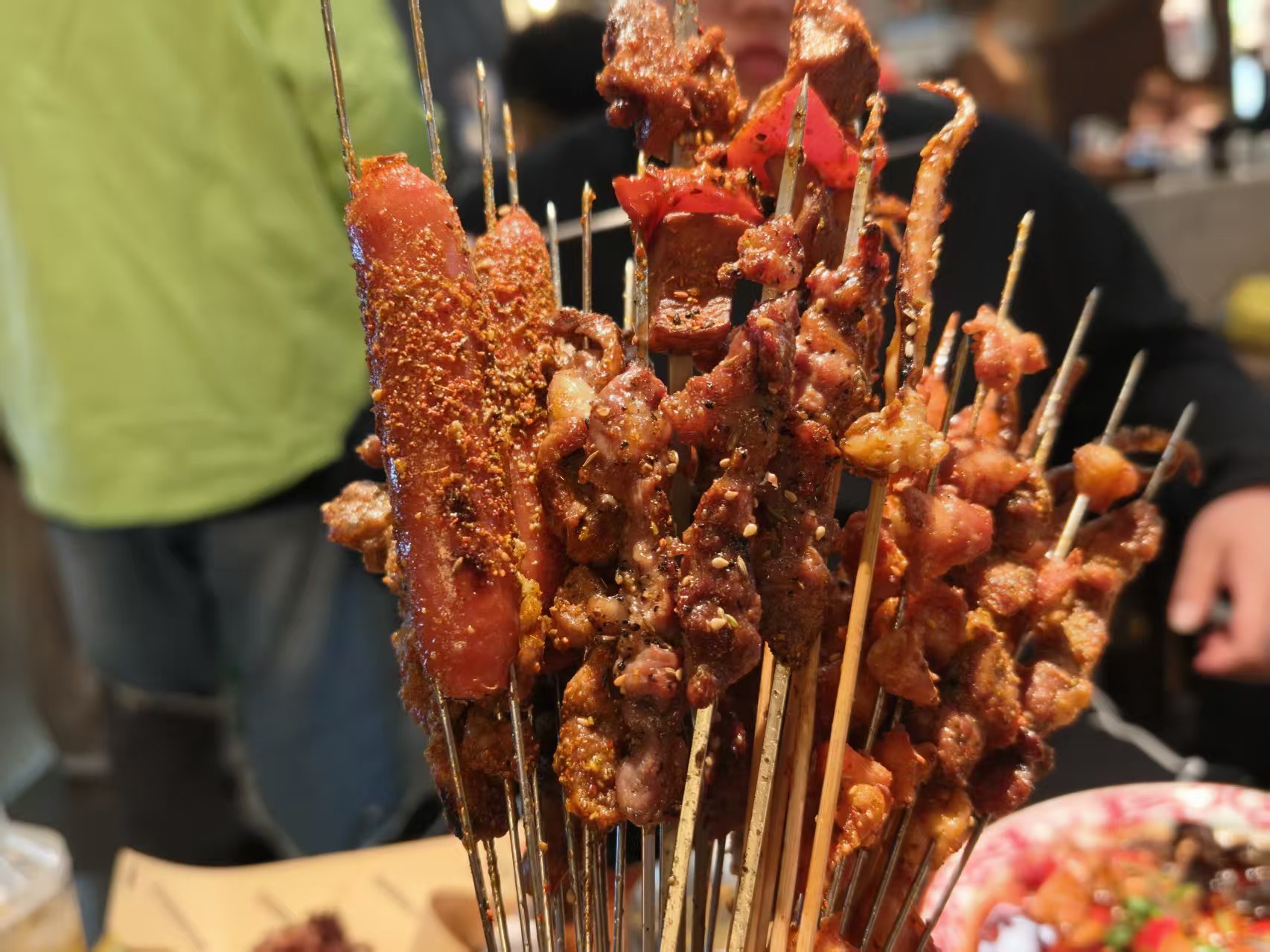 堆叠在一起的烧烤串,肉串、香肠串、鱿鱼串,表面撒芝麻和辣椒面