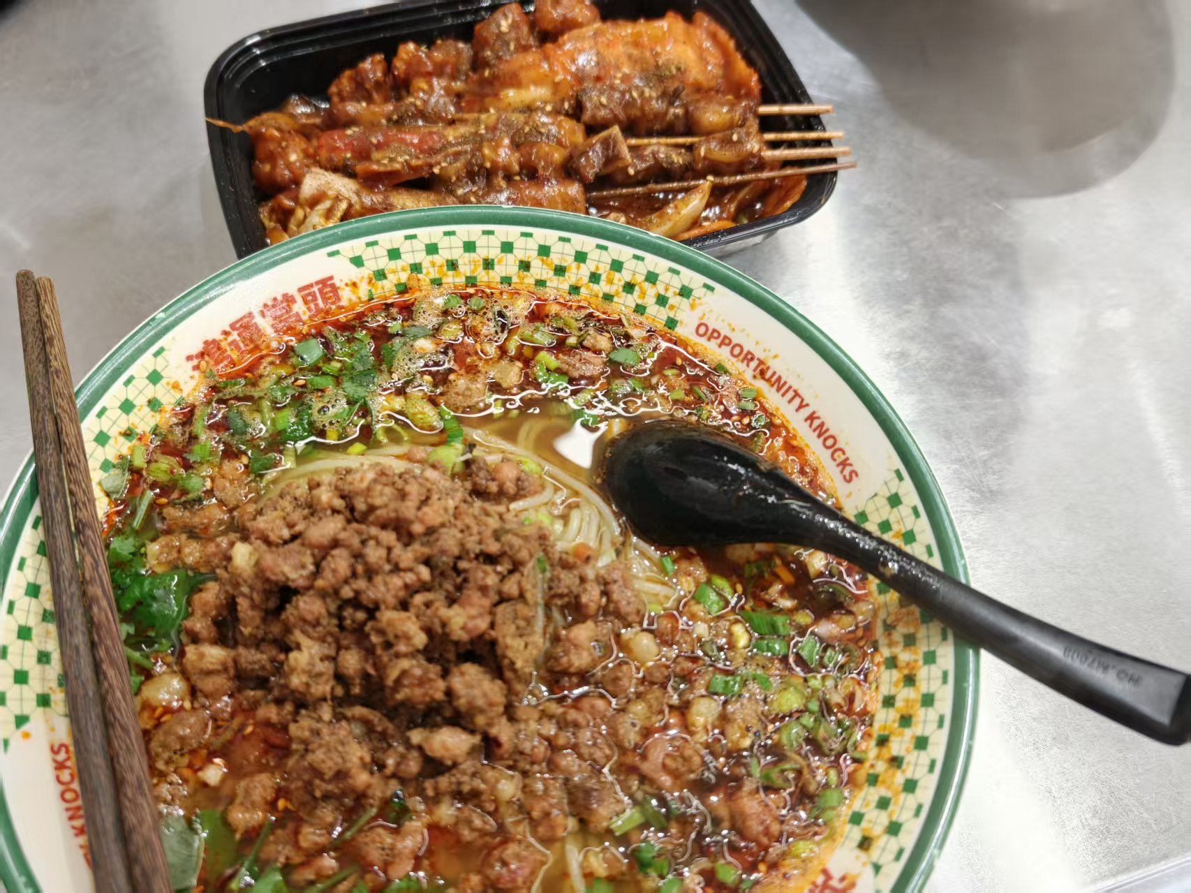 红油汤底的杂酱米粉,碗身印鸿运当头,旁边黑餐盒里是串在竹签上的烤串