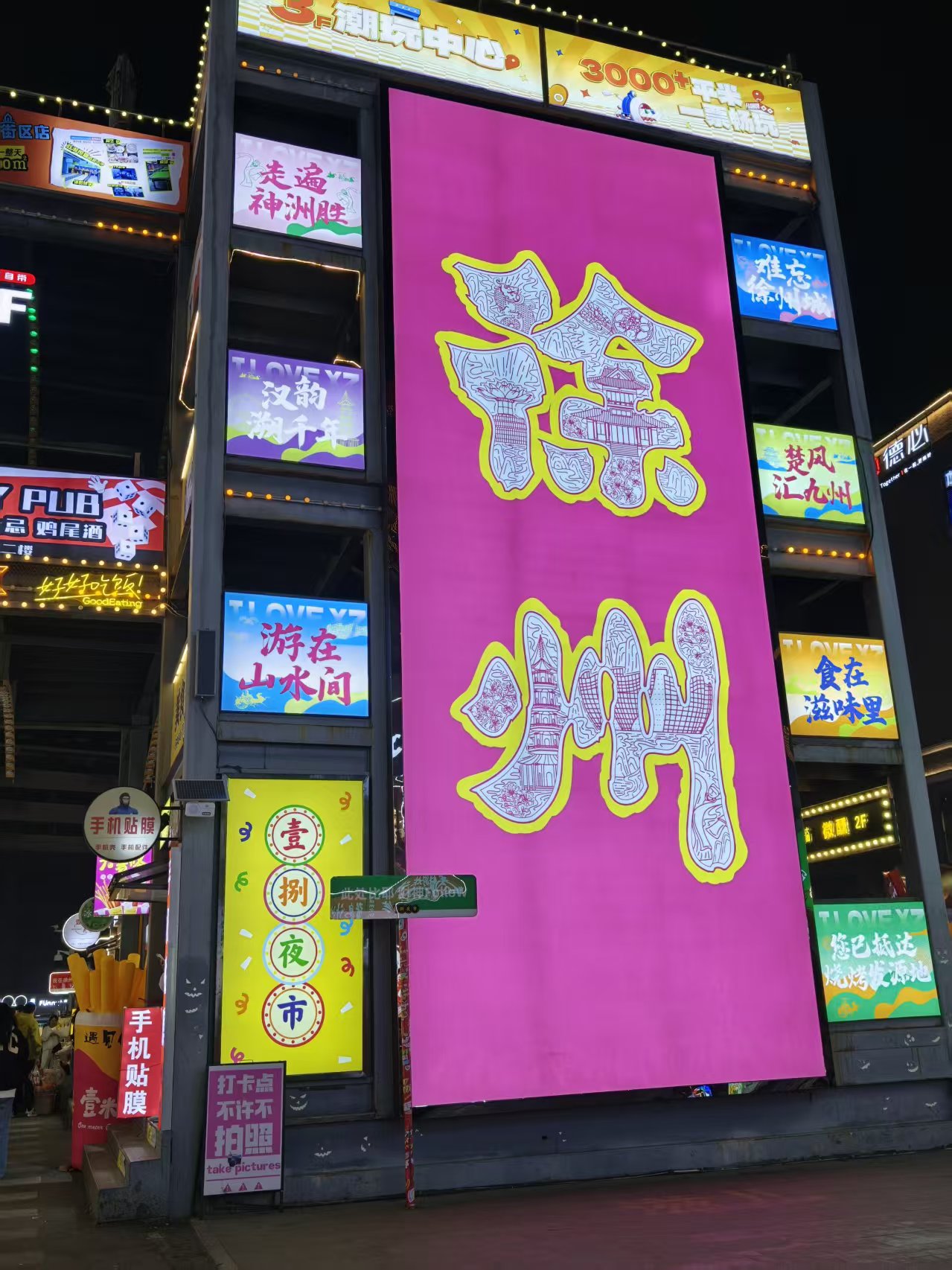夜晚商业街区外立面,巨大玫红竖幅广告牌写徐州二字,四周是潮玩、夜市、餐饮招牌,灯带亮起